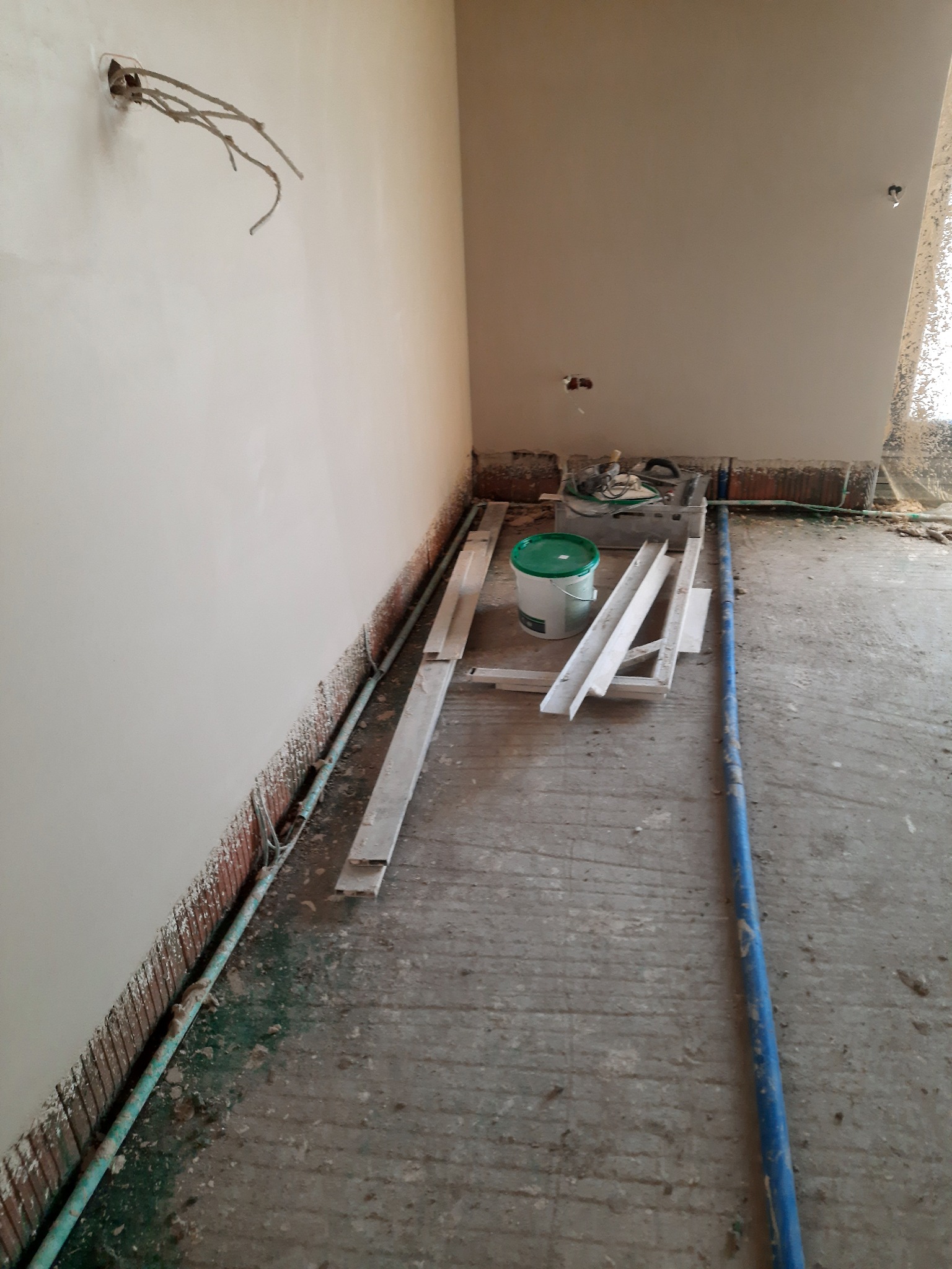 Świeżo otynkowane ściany w rogu pomieszczenia, widoczne przewody elektryczne wystające ze ściany, wiadro z zieloną pokrywką i elementy rusztowania na podłodze w trakcie prac wykończeniowych.