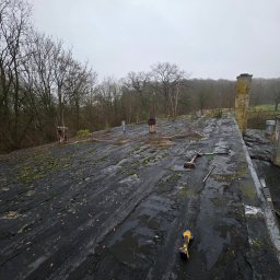 Dachy STEFANIAK - Demontaż starego, zniszczonego dachu pokrytego mchem. Widoczne narzędzia pracy i fragment komina. W tle drzewa i pochmurne niebo.