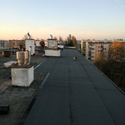 Dachy STEFANIAK - Wykończony dach pokryty papą termozgrzewalną w Wałczu. Widoczne kominy i pracownik z narzędziami. W tle zabudowa miejska i niebo o zachodzie słońca.