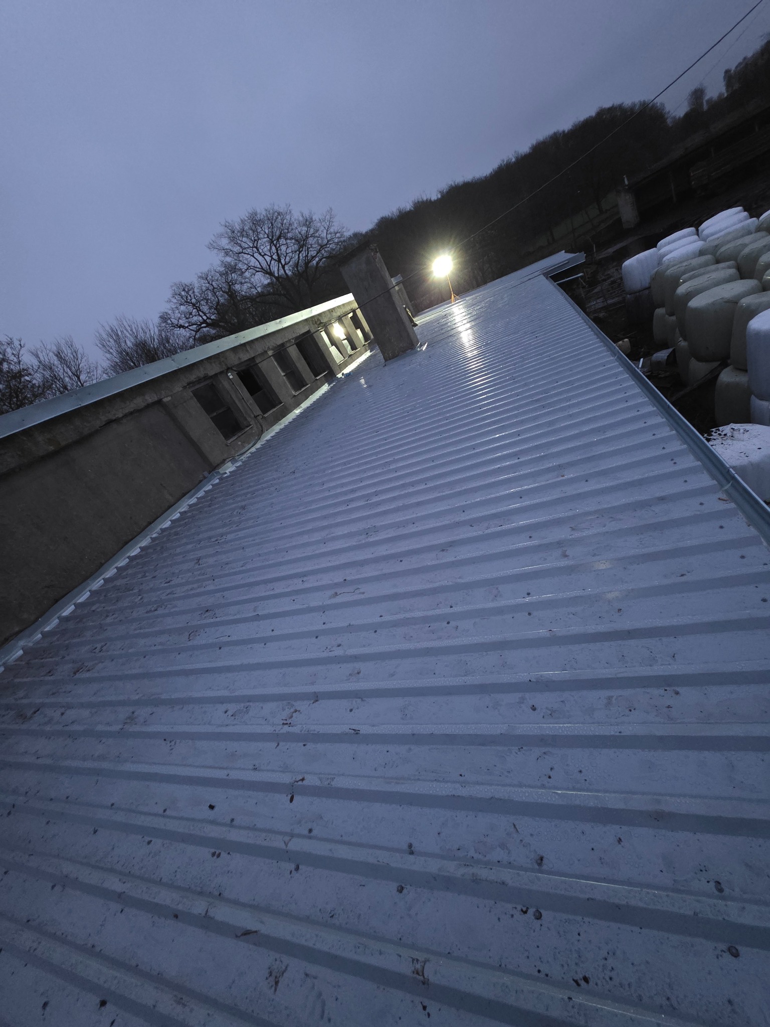 Nowy, srebrny dach z blachy trapezowej, oświetlony lampą, widoczny pod kątem. W tle drzewa i białe bele siana. Widoczne okna w budynku.