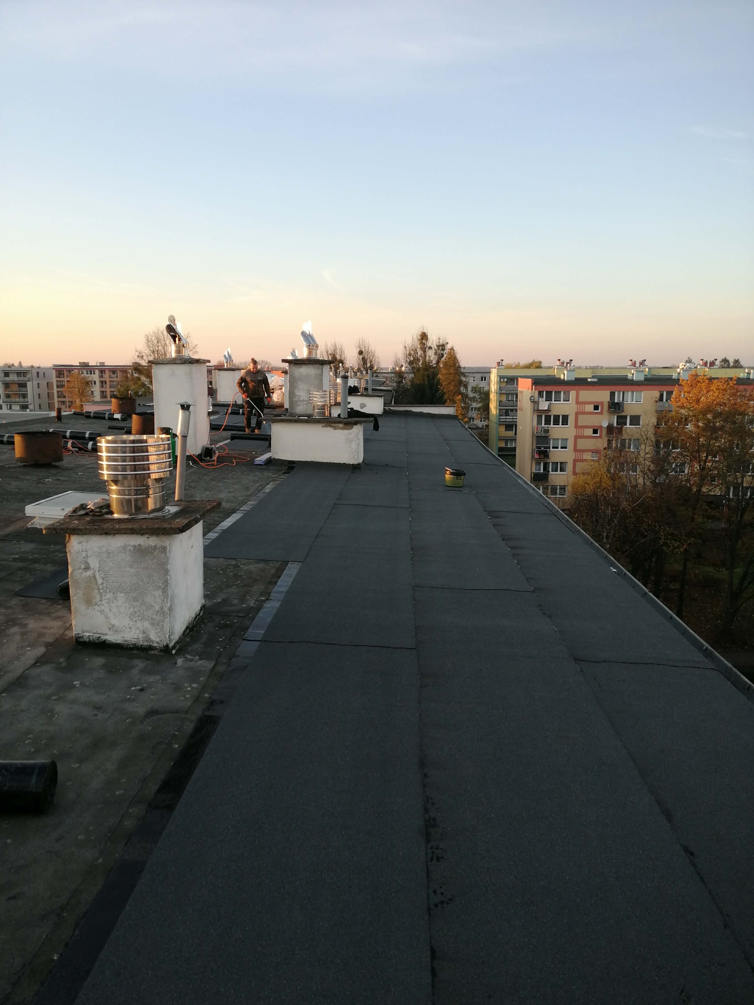 Wykończony dach pokryty papą termozgrzewalną w Wałczu. Widoczne kominy i pracownik z narzędziami. W tle zabudowa miejska i niebo o zachodzie słońca.