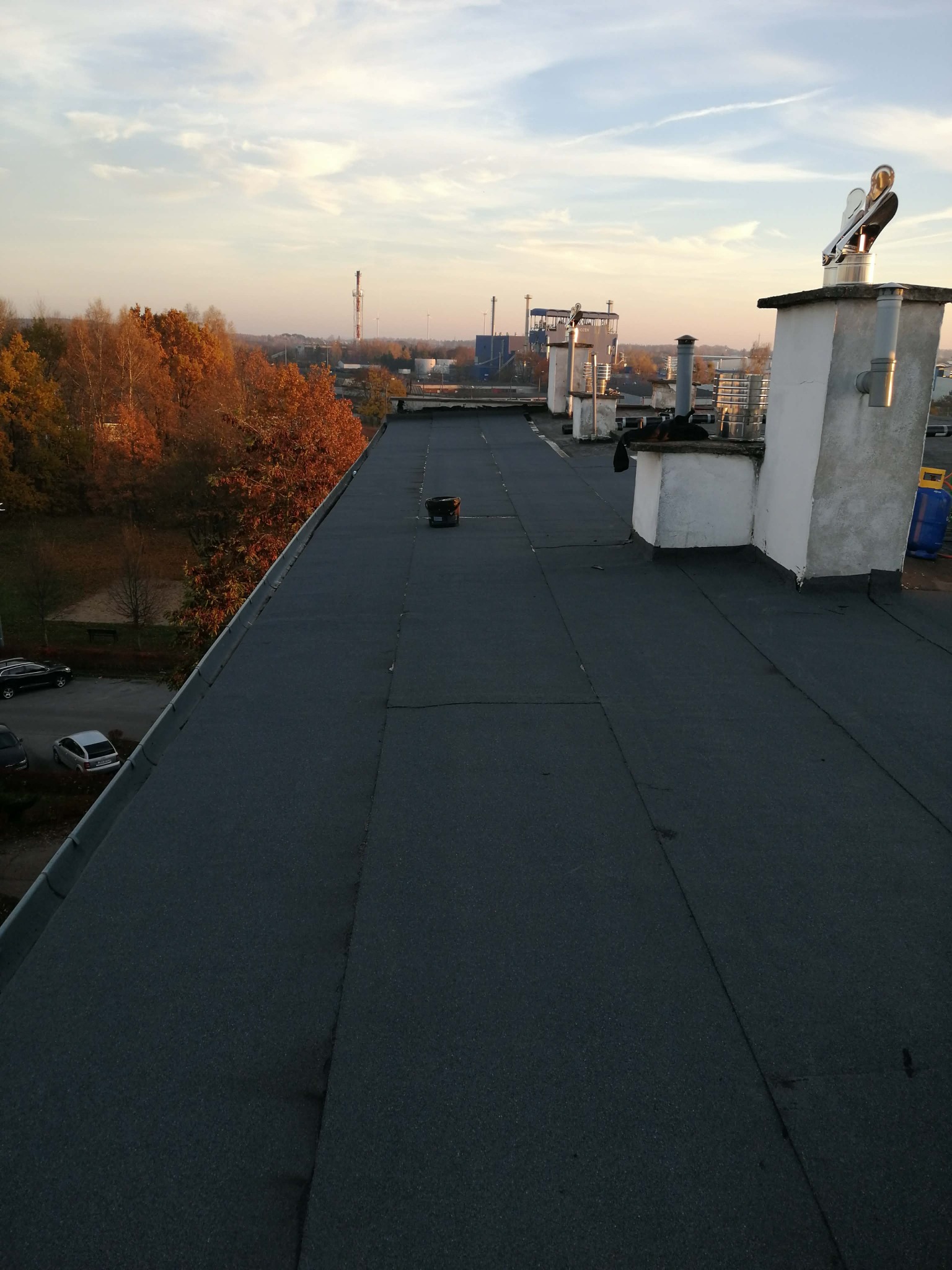 Czarny, płaski dach budynku z kominami wentylacyjnymi i widokiem na jesienne drzewa oraz zabudowania przemysłowe w oddali, sfotografowany z góry.