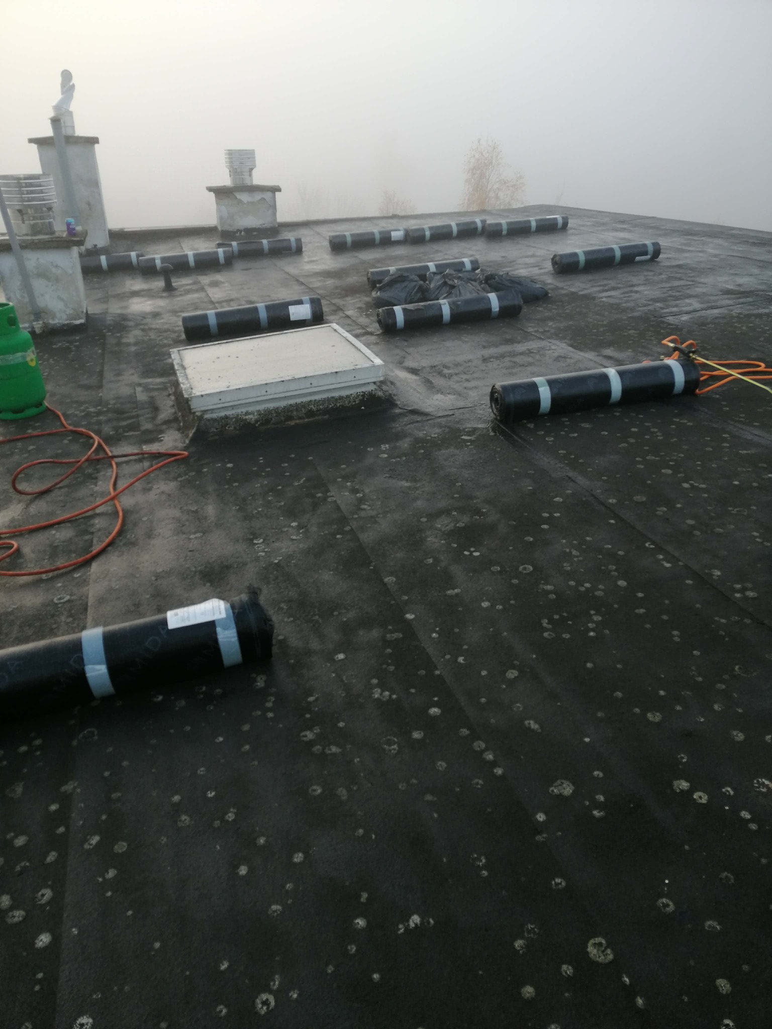 Czarny dach pokryty papą z widocznymi rolkami papy przygotowanymi do montażu, butla gazowa i przewody. W tle kominy i mgła.