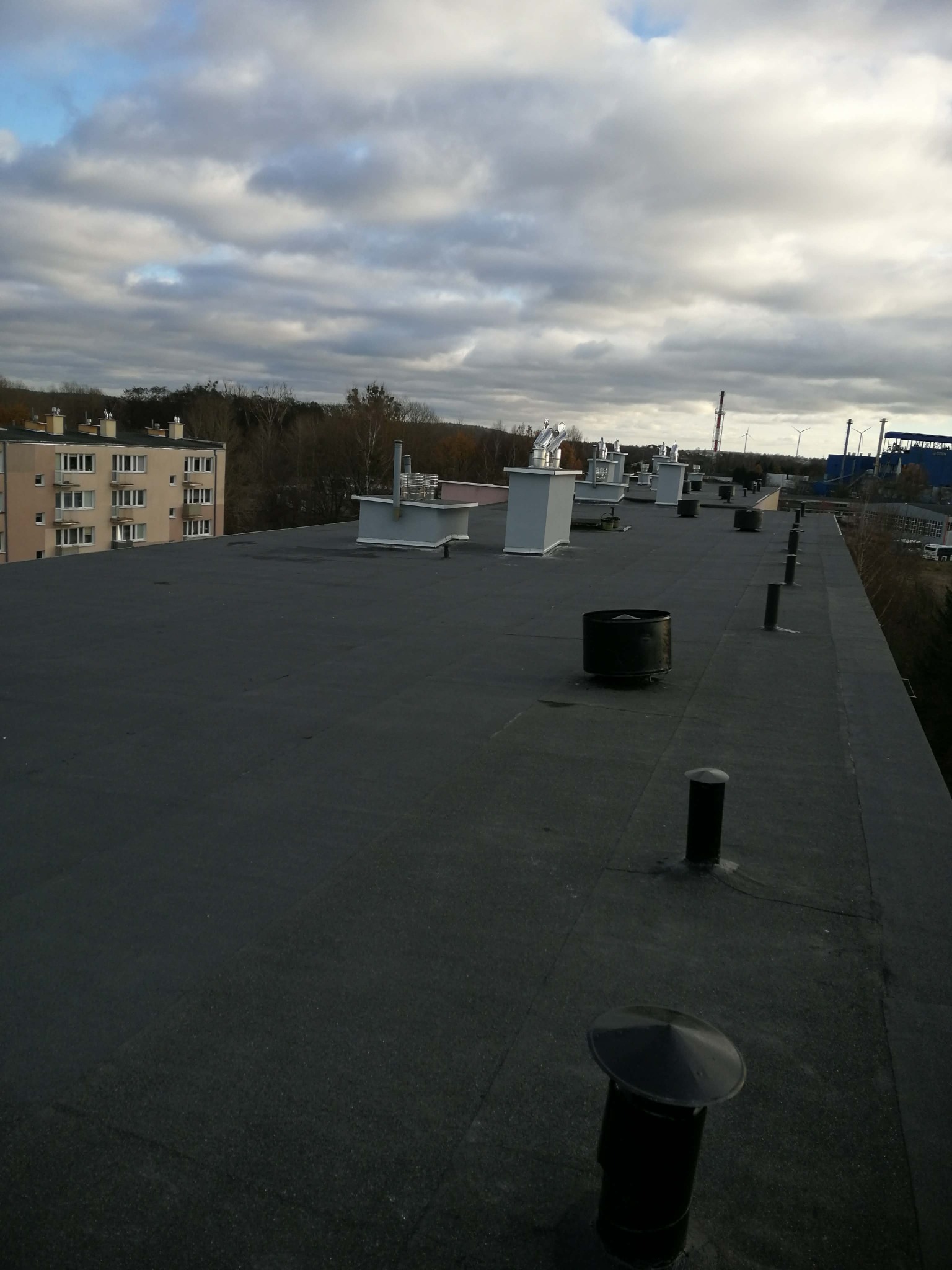Płaski dach budynku z kominami wentylacyjnymi i odpowietrznikami na tle pochmurnego nieba i zabudowań miejskich. Widok z góry, perspektywa z boku.