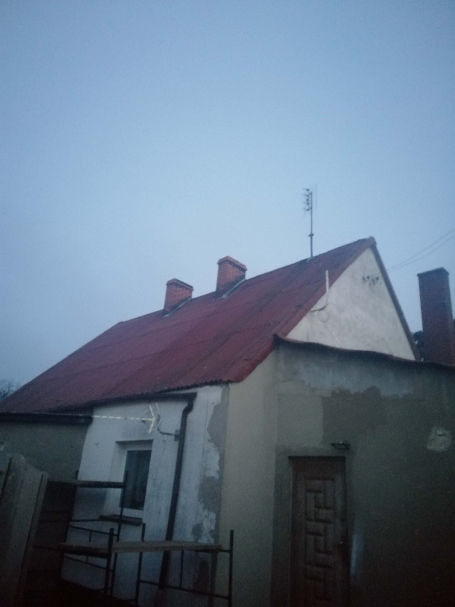 Dom z czerwonym dachem i kominami, widoczny z dołu pod kątem. Ściany częściowo otynkowane, z widoczną anteną na dachu. Pochmurne niebo w tle.