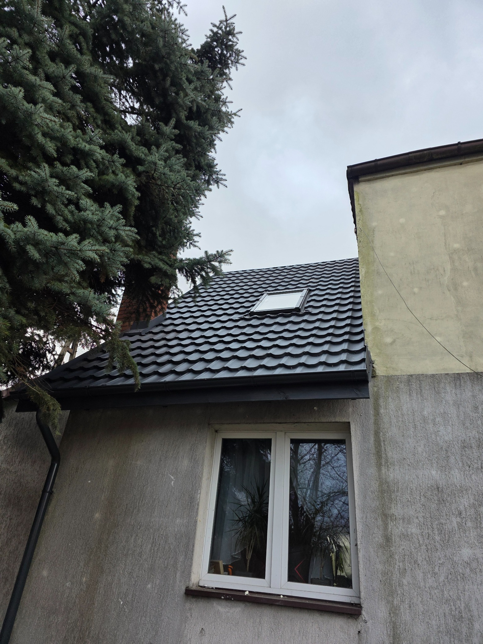 Dom z nowym, ciemnym dachem z oknem dachowym, widoczny z dołu, obok iglaste drzewo. Szara elewacja i okno z odbiciem drzewa.
