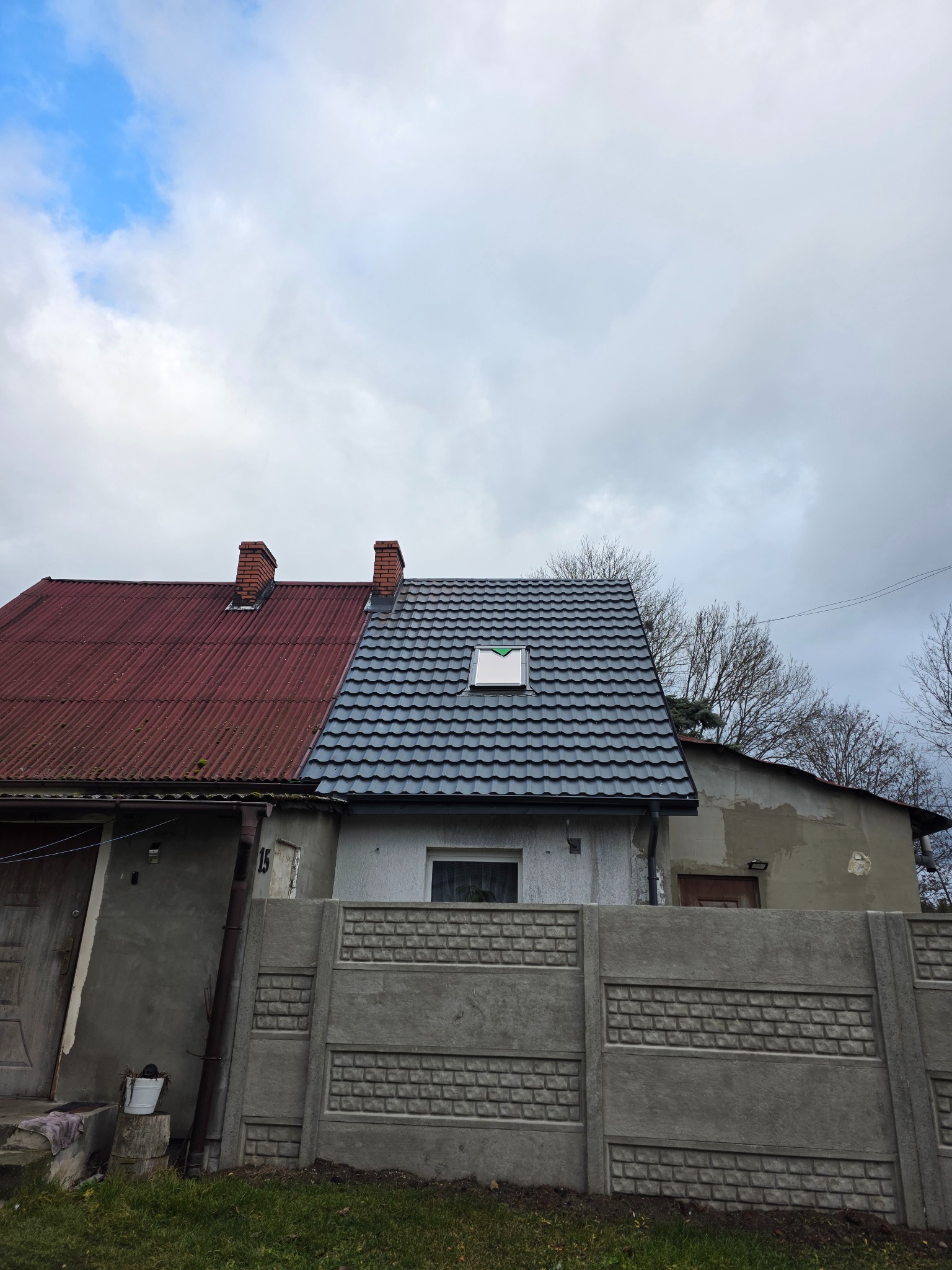 Dom z nowym, grafitowym dachem z blachy trapezowej i oknem dachowym, kontrastujący z sąsiednim budynkiem o czerwonym dachu. Betonowe ogrodzenie na pierwszym planie.