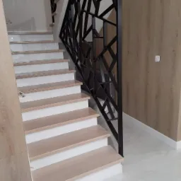 Nowoczesne schody z jasnego drewna z białymi podstopnicami i geometryczną, czarną balustradą w minimalistycznym wnętrzu z panelami ściennymi imitującymi drewno.