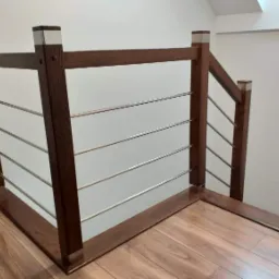 Fragment drewnianej balustrady z poziomymi, metalowymi prętami, wykończonej srebrnymi akcentami, na tle białej ściany i drewnianej podłogi.