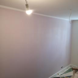 Wykończeni - Świeżo pomalowana na liliowo ściana z widoczną listwą przypodłogową i prowizorycznym oświetleniem w postaci żarówki na kablu, podłoga częściowo zabezpieczona dywanem dziecięcym.