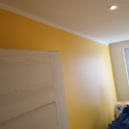 Świeżo pomalowana na żółto ściana z zamontowaną drewnianą ościeżnicą i drzwiami w jasnym odcieniu drewna, widoczny sufit z oświetleniem punktowym i fragment okna z roletą.