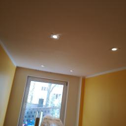 Wykończeni - Pokój po remoncie z pomalowanymi na żółto ścianami, białym sufitem z zamontowanymi punktowymi lampami LED i oknem z widokiem na zewnątrz, z widocznymi materiałami budowlanymi przy oknie.