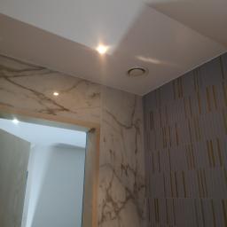 Wykończeni - Wykończona łazienka z marmurowymi płytkami imitującymi wzór żyłkowania na ścianie obok wejścia, oraz szare płytki z pionowymi złotymi paskami na sąsiedniej ścianie, widoczny fragment sufitu...