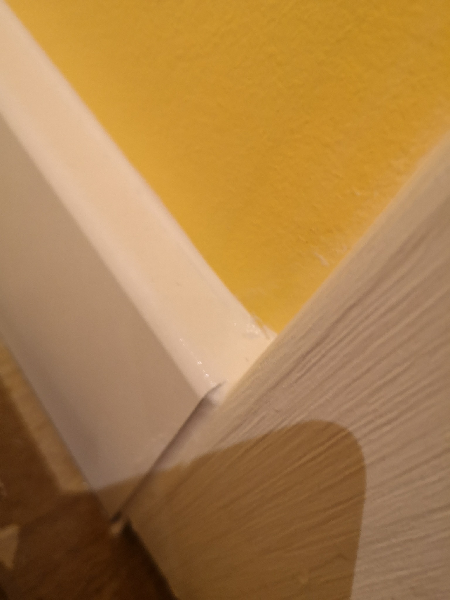 Szczegółowe ujęcie łączenia białej listwy przypodłogowej z żółtą ścianą i drewnianą podłogą, widoczne niedociągnięcia w malowaniu.