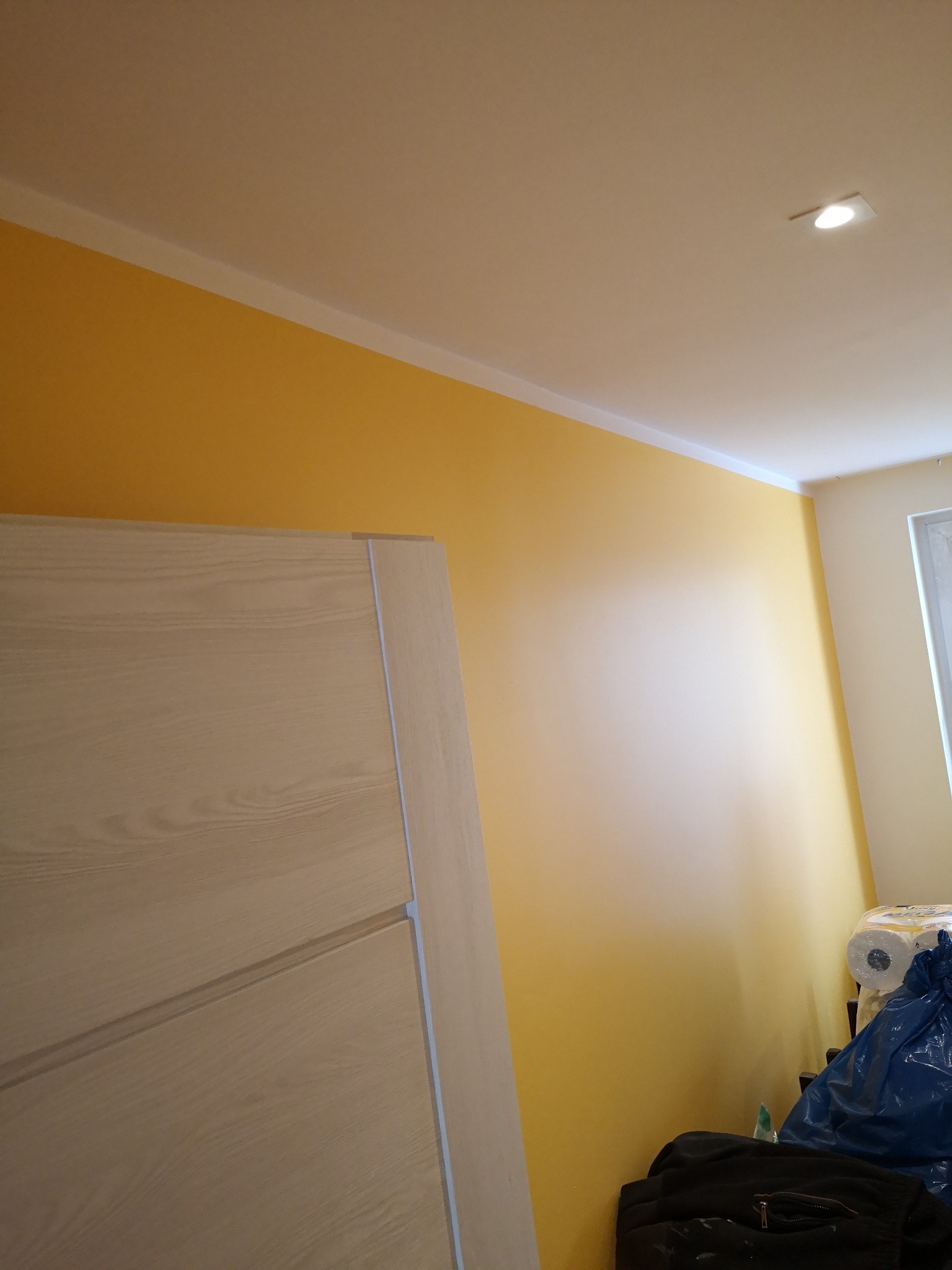 Świeżo pomalowana na żółto ściana z zamontowaną drewnianą ościeżnicą i drzwiami w jasnym odcieniu drewna, widoczny sufit z oświetleniem punktowym i fragment okna z roletą.