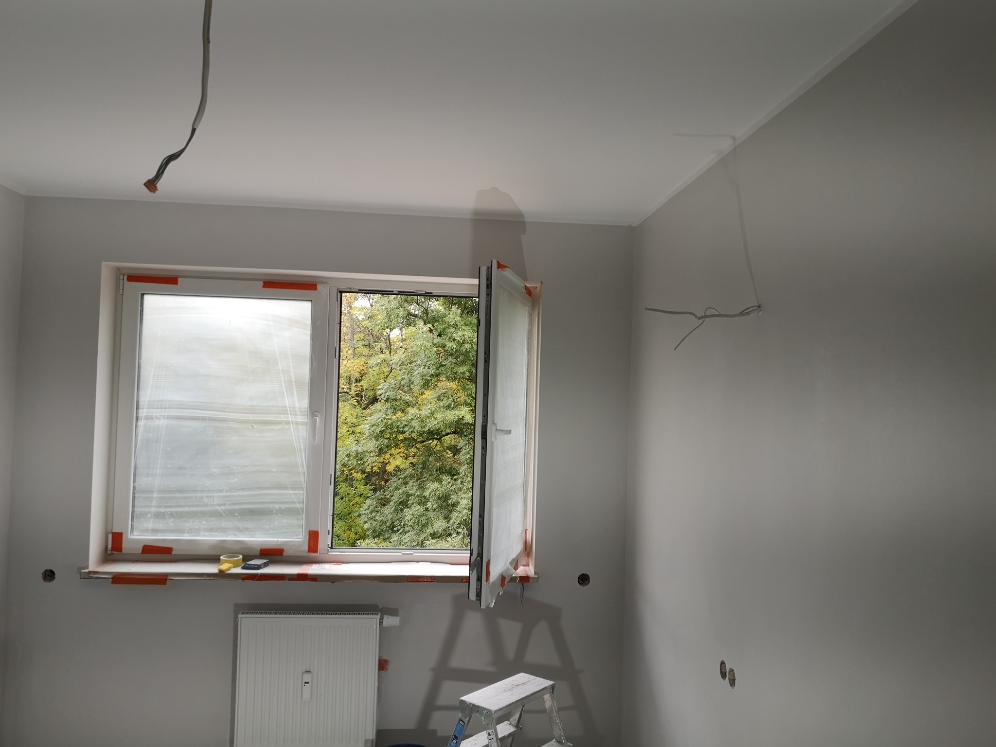 Pokój w trakcie odświeżania: ściany pomalowane na szaro, okno zabezpieczone folią i taśmą malarską, widoczny grzejnik i drabina malarska, wystające kable elektryczne.