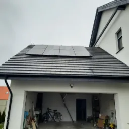 Panele słoneczne zamontowane na dachu domu z garażem, widok z poziomu gruntu, pochmurne niebo w tle.