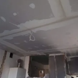 Niedokończona powierzchnia sufitu z widocznymi śladami po nałożeniu pierwszej warstwy masy szpachlowej, wystający kabel oświetleniowy, fragment ściany z piecem gazowym w tle.