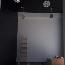 Wnętrze w trakcie prac: ściana pomalowana na szaro i czarno, drewniana drabina oparta o ścianę, widoczne lampy sufitowe i narzędzia malarskie na podłodze.