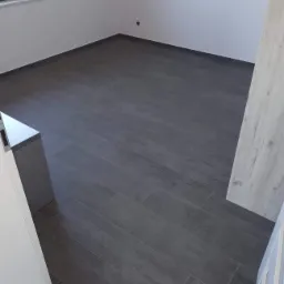 Ułożona, ciemnoszara podłoga z płytek imitujących drewno w pustym pomieszczeniu z białymi ścianami, widok z perspektywy wejścia.