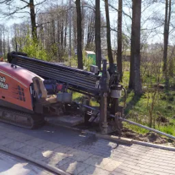 Pomarańczowa wiertnica Ditch Witch na chodniku z rurami wiertniczymi, z widoczną łopatą i znakiem przejścia dla pieszych w tle, na tle drzew.