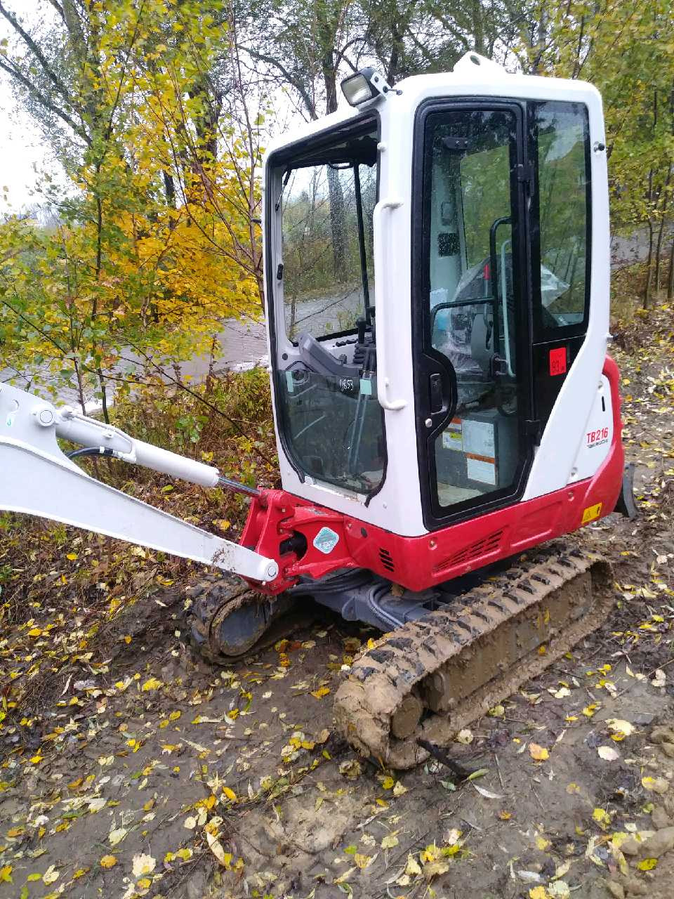 Mała, biało-czerwona koparka gąsienicowa na błotnistym terenie z opadłymi liśćmi, widoczna kabina operatora z naklejką 0853 na szybie.