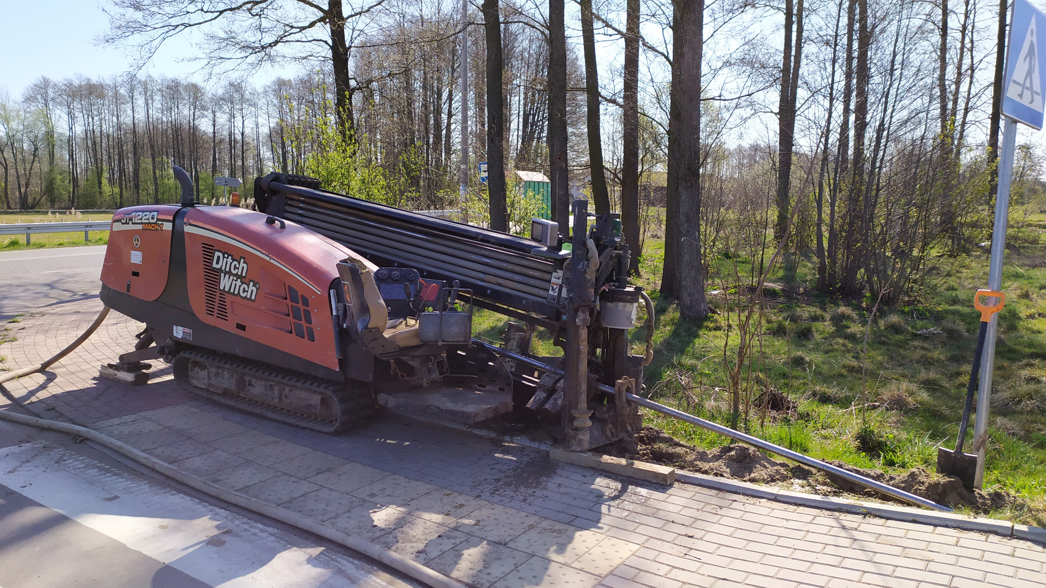 Pomarańczowa wiertnica Ditch Witch na chodniku z rurami wiertniczymi, z widoczną łopatą i znakiem przejścia dla pieszych w tle, na tle drzew.