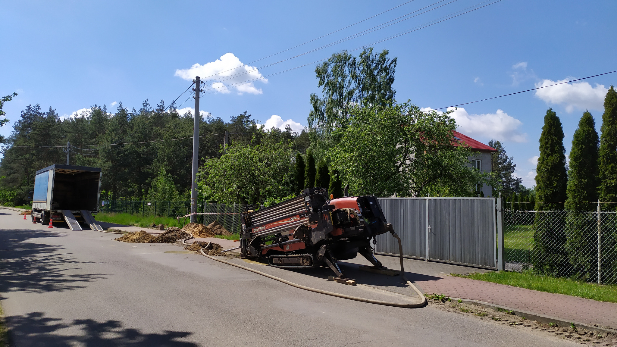 Wiertnica horyzontalna Ditch Witch JT2020 na asfalcie, z widocznym transportowym samochodem ciężarowym i wykopem w tle, podczas prac instalacyjnych w słoneczny dzień.