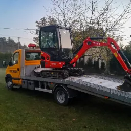 Żółty samochód pomocy drogowej z czerwonym minikoparką Kubota KX018-4 na platformie transportowej, na tle zielonego trawnika i drzew bez liści.