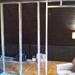 Łukasz Budownictwo - Metalowa konstrukcja szkieletowa ścianki działowej w pokoju na poddaszu z żółtą ścianą i brązową boazerią, widoczne narzędzia i kartonowe pudło.