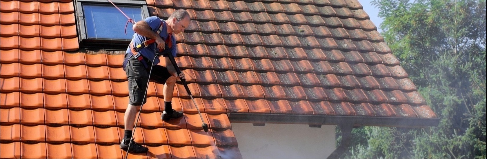Pracownik w uprzęży, myjący ciśnieniowo dach z czerwonej dachówki, widoczne zabrudzenia na części dachu przed czyszczeniem, okno dachowe w tle.