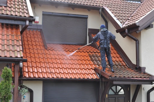 Pracownik w kombinezonie zabezpieczającym czyści dach z czerwonej dachówki za pomocą myjki ciśnieniowej, widoczne zacieki i różnica w czystości powierzchni.