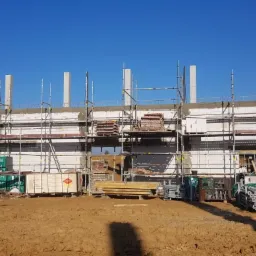 Budowa budynku w stanie surowym otwartym, widoczne szare słupy konstrukcyjne, białe ściany z pustaków, rusztowania, materiały budowlane i ziemia.