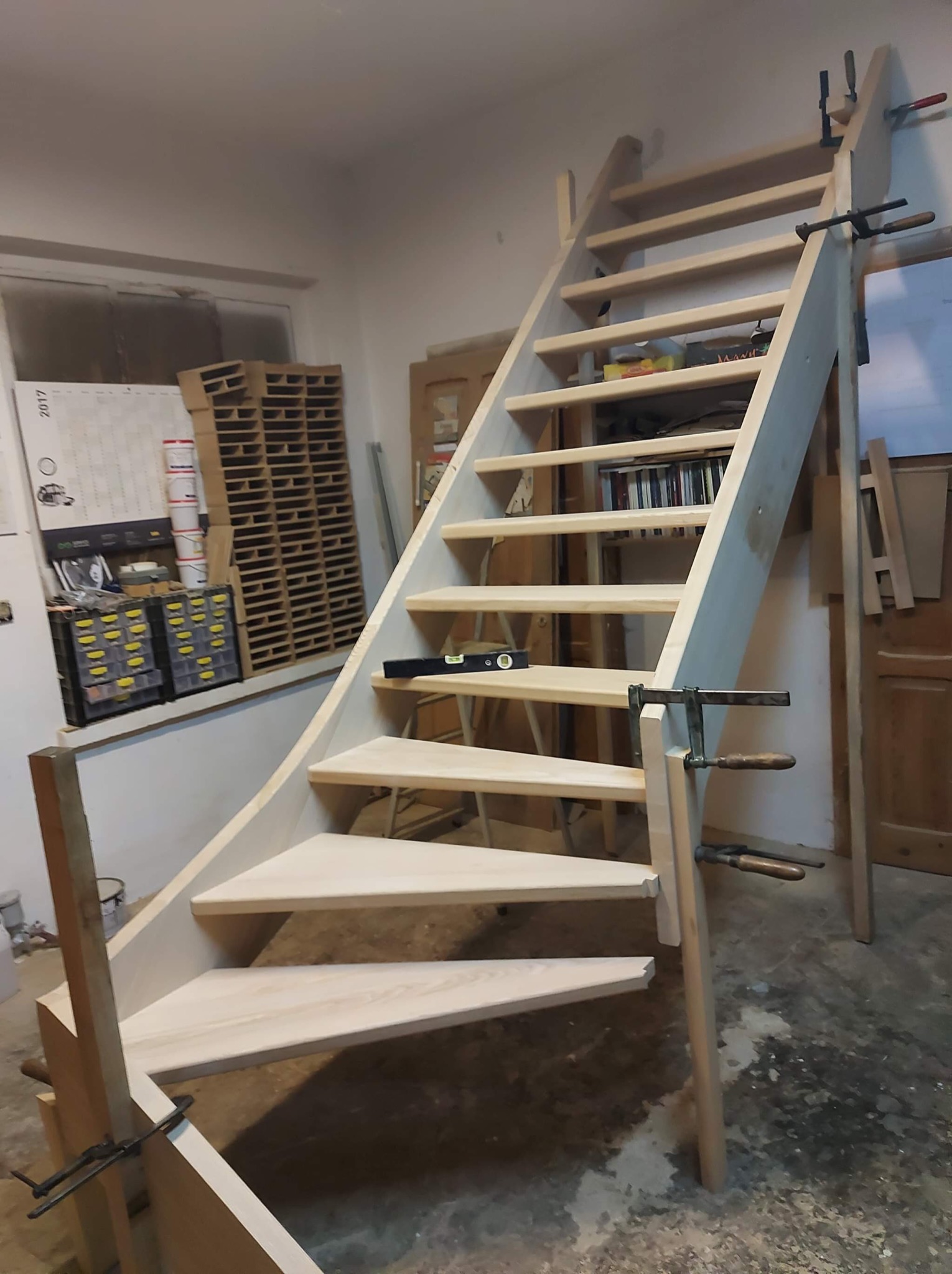 Drewniane schody z jasnego drewna w trakcie montażu, zabezpieczone ściskami stolarskimi, widoczny poziomica na jednym ze stopni, w tle okno i regał z pojemnikami.