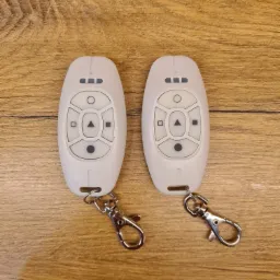 Dwa białe piloty do systemu sterowania na drewnianym blacie, z metalowymi karabińczykami, widoczne przyciski funkcyjne.