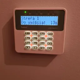 Różowy panel sterowania systemu alarmowego Satel Perfecta, zamontowany na ścianie, wyświetlający komunikat 'Strefa 1, Do wyjścia: 13s'.