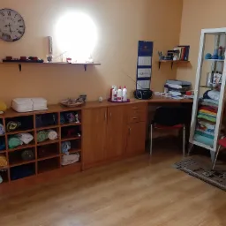 Pokój zabiegowy z szafką pełną zwiniętych ręczników, kosmetykami na półce i zegarem na ścianie.