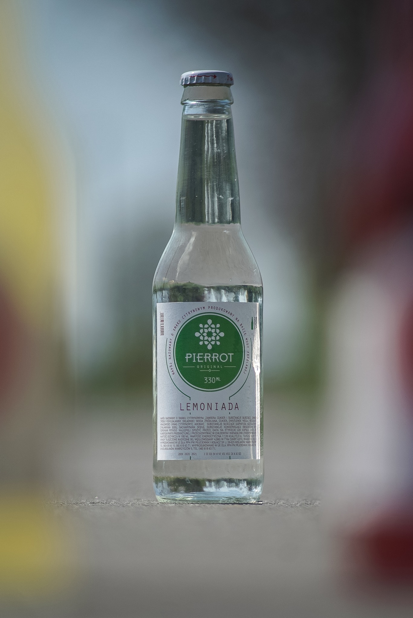 Butelka lemoniady Pierrot Original 330ml, ustawiona na szarej powierzchni, z rozmytym tłem w odcieniach zieleni i czerwieni.