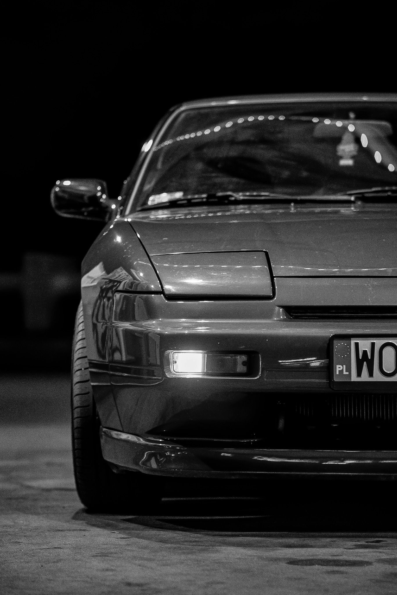 Czarno-białe ujęcie z bliska przedniej części sportowego samochodu z włączonym światłem pozycyjnym i polską tablicą rejestracyjną.