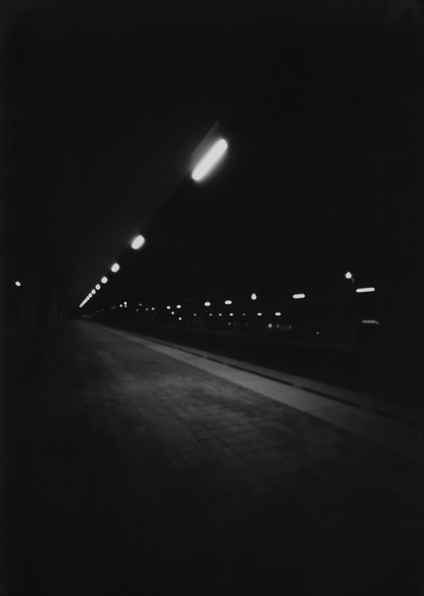 Czarno-białe zdjęcie wykonane techniką otworkową przedstawiające oświetlony tunel lub peron metra z perspektywy podłoża.