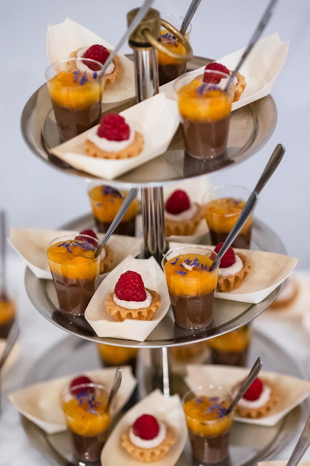 Elegancki, trzypoziomowy etażer z deserami: mini tartaletki z maliną i kremem oraz czekoladowe musy z musem mango i płatkami kwiatów, każdy deser na papierowej łódeczce, srebrne łyżeczki w każdym...