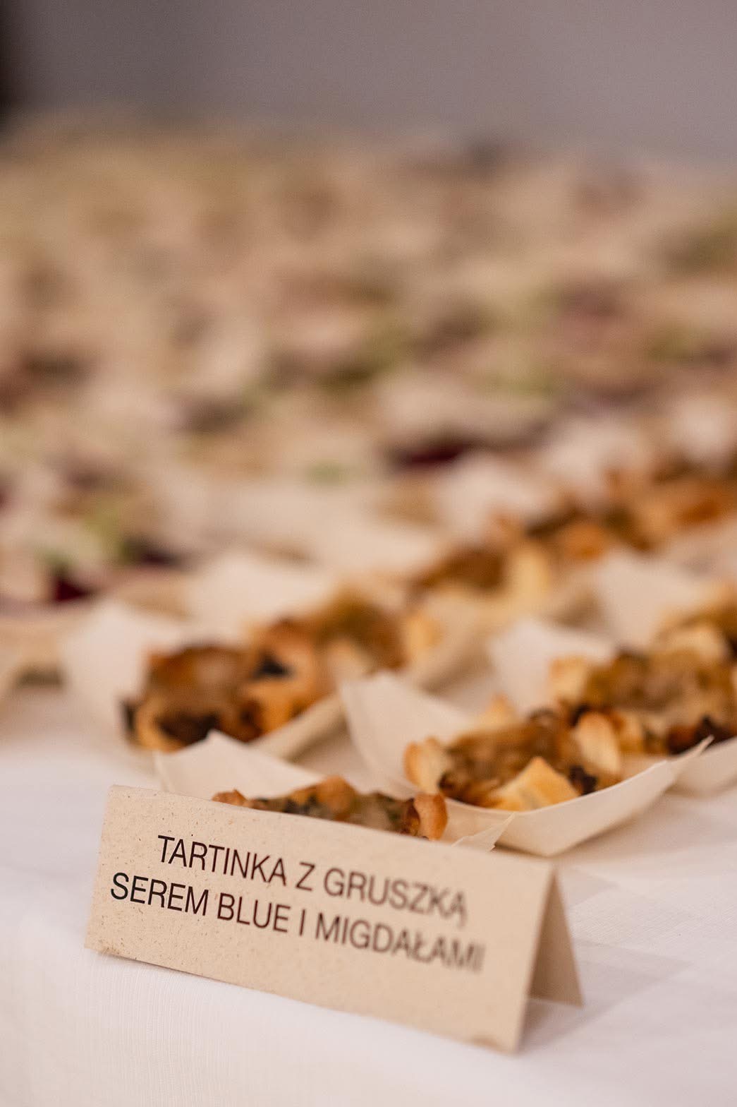 Rząd tartinek z gruszką, serem blue i migdałami, podanych na białym stole w małych, ekologicznych miseczkach, z widoczną etykietą z nazwą deseru.