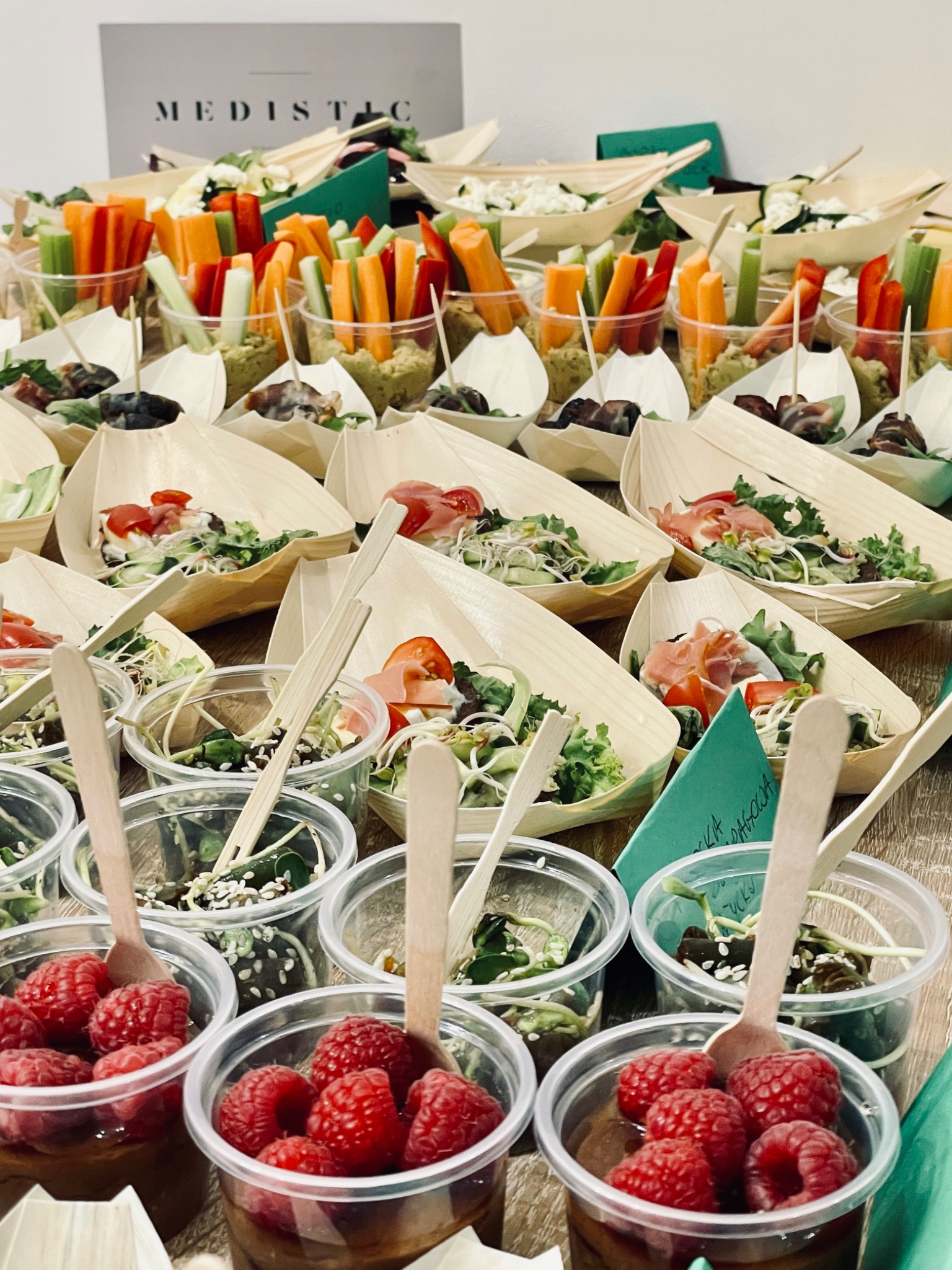 Różnorodność przekąsek cateringowych: mini szaszłyki z warzywami, sałatki w łódeczkach z jasnego drewna i desery z malinami w przezroczystych kubeczkach, z drewnianymi łyżeczkami, na stole podczas...