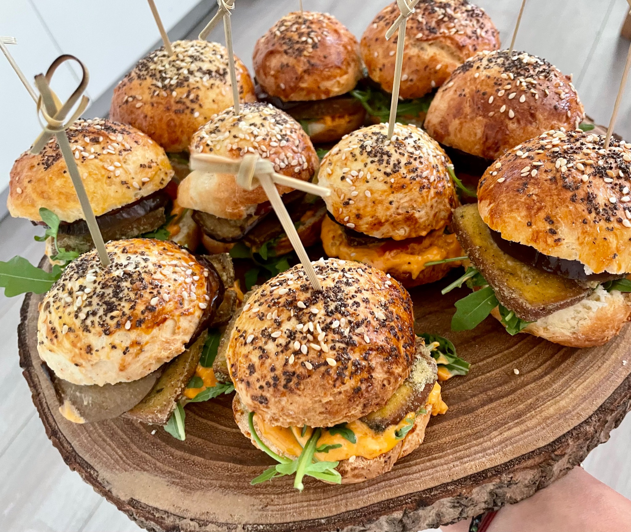 Mini burgery z grillowanym bakłażanem, rukolą i sosem pomarańczowym, spięte wykałaczkami, ułożone na drewnianej desce imitującej plaster drzewa.
