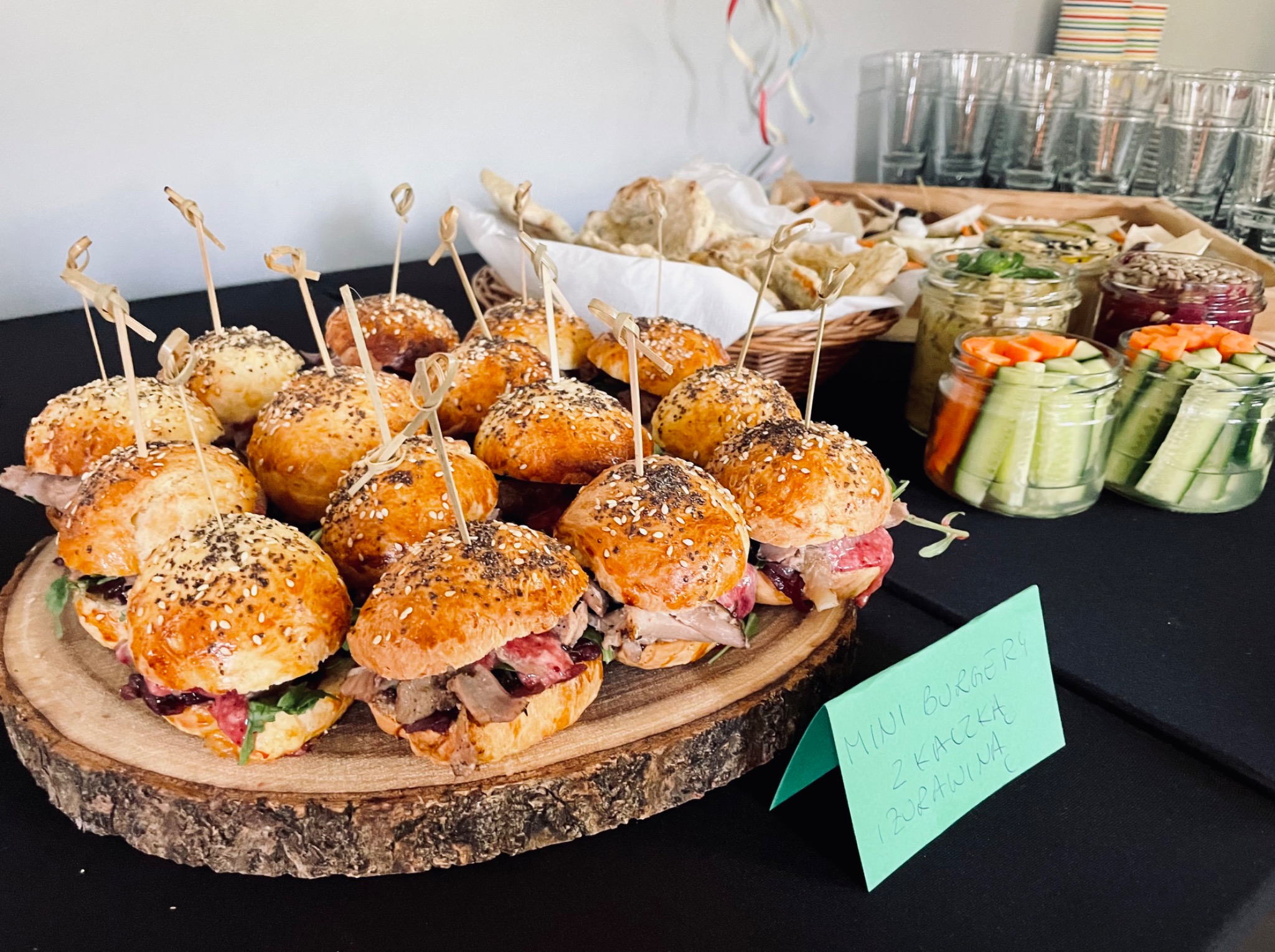 Mini burgery z kaczką i żurawiną na drewnianej desce, przekąski w słoikach i stos szklanek w tle, przygotowane na przyjęcie.