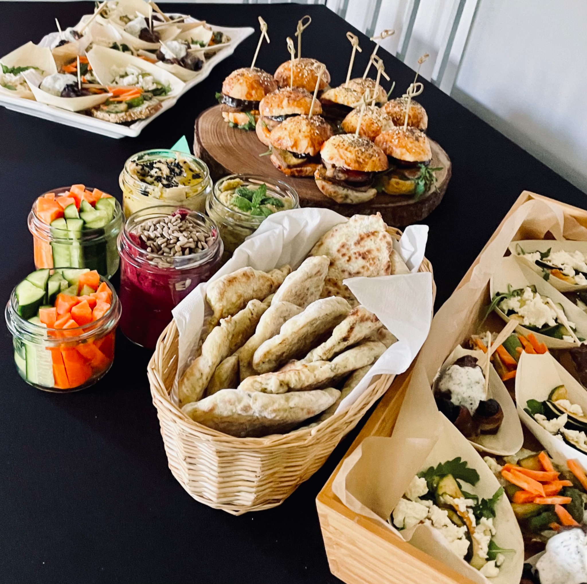 Bogaty wybór przekąsek na czarnym stole: mini burgery na drewnianym plastrze, pieczywo w wiklinowym koszyku, słoiczki z pokrojonymi warzywami i pastami, oraz porcje sałatek w drewnianych łódeczkach.