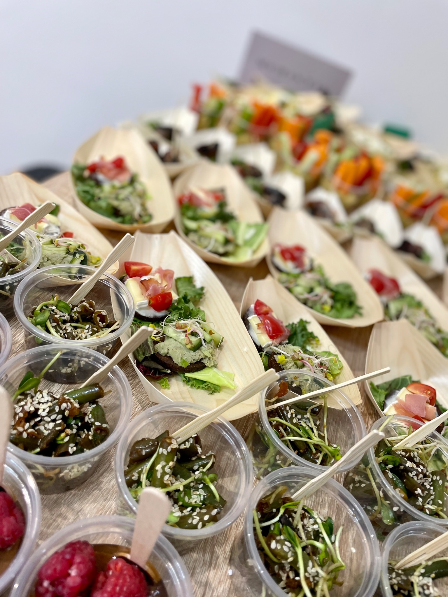 Różnorodne przekąski w małych, przezroczystych miseczkach i drewnianych łódeczkach, ułożone rzędami na jasnym stole. Widoczne sałatki, warzywa, owoce i zioła, niektóre z dodatkiem sezamu, podane...