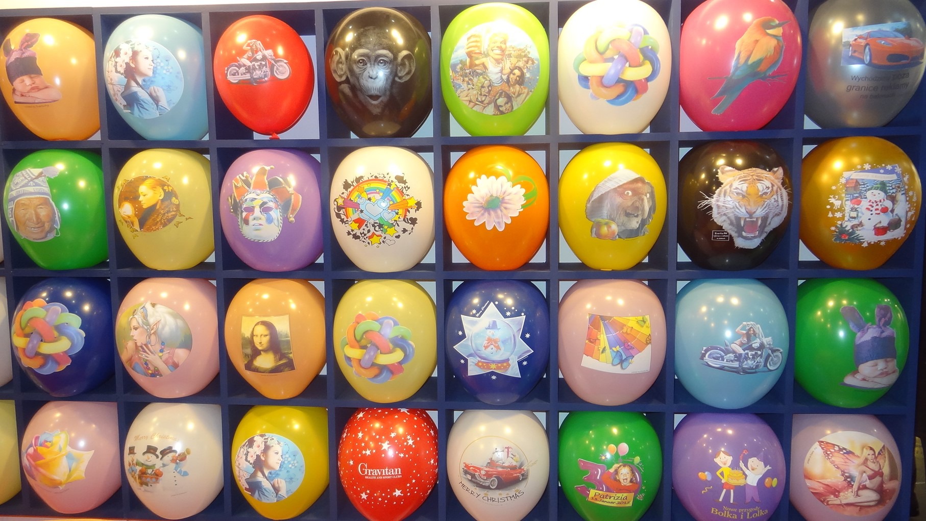Różnokolorowe balony z nadrukami różnych motywów, umieszczone w niebieskim regale z przegródkami, tworzące barwną kompozycję.