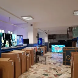 Magazyn z zapakowanymi telewizorami różnych marek, niektóre ekrany włączone, oświetlenie sufitowe LED.