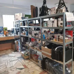 Półki magazynowe wypełnione różnorodnym sprzętem elektronicznym, w tym kamerami, stabilizatorami, drukarkami i akcesoriami w oryginalnych opakowaniach, na tle wzorzystej posadzki.